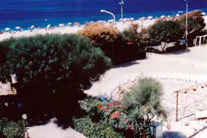 Blick auf die Strandpromenade mit weißen Sonnenschirmen und Liegen am Meer, umgeben von Bäumen und Hecken.