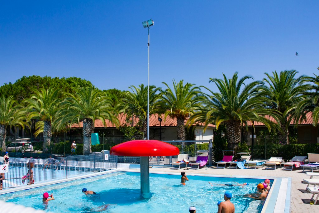 Kinder und Erwachsene im Schwimmbecken am Campingplatz, umgeben von Palmen, Liegen und Sonnenschirmen.