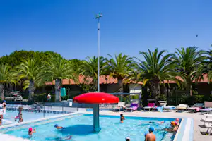 Kinder und Erwachsene im Schwimmbecken am Campingplatz, umgeben von Palmen, Liegen und Sonnenschirmen.