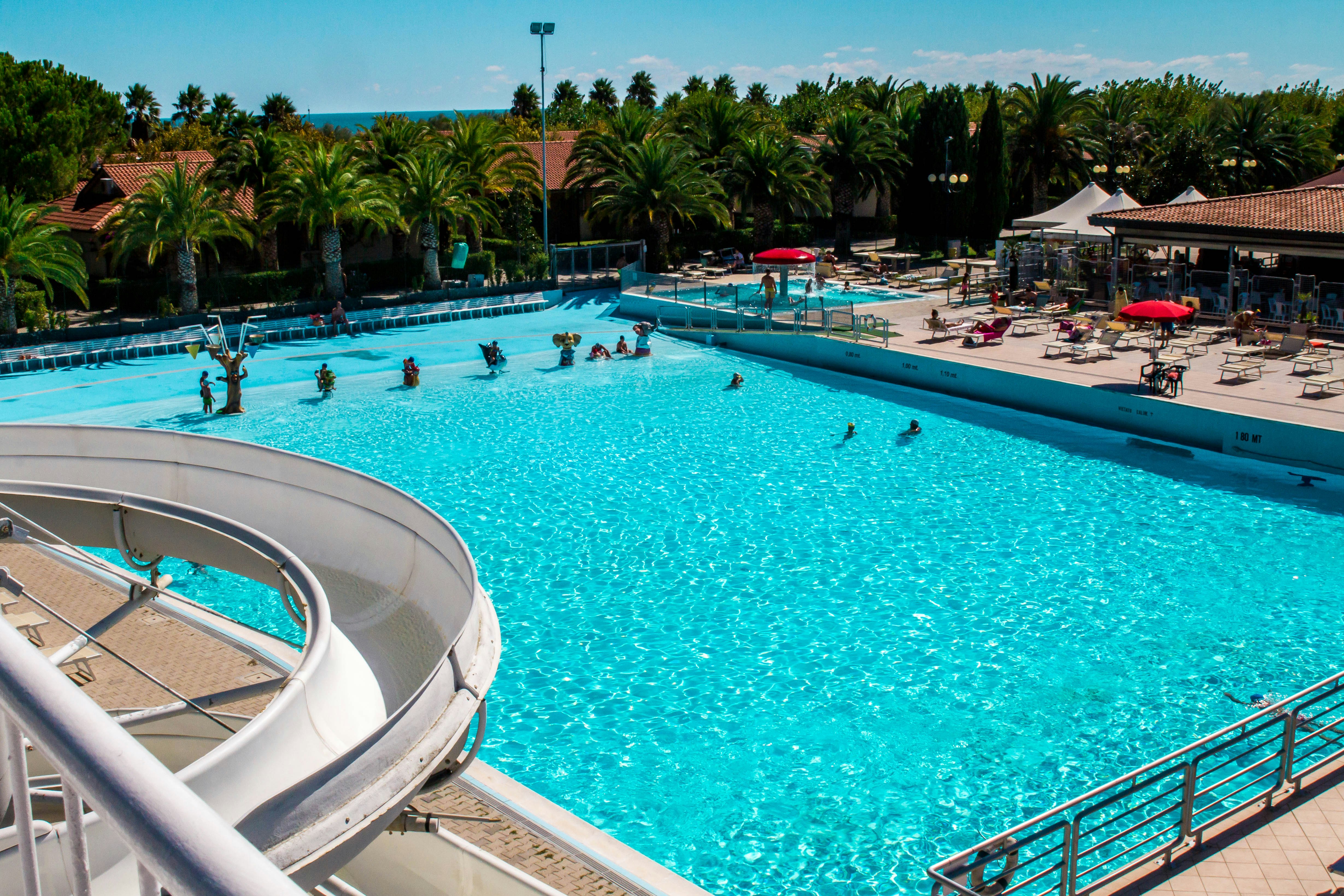 Großes Camping-Schwimmbecken mit Treppe, Liegen und Sonnenschirmen, im Hintergrund Palmen und Gebäude am Poolrand.