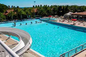 Großes Camping-Schwimmbecken mit Treppe, Liegen und Sonnenschirmen, im Hintergrund Palmen und Gebäude am Poolrand.