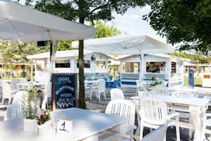 Terrasse mit weißen Tischen und Stühlen unter Sonnenschirmen, im Hintergrund weitere Sitzbereiche und Bäume.