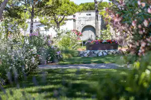Blumenbeete und Rasenfläche im Campinggarten mit Steinbrunnen und Weg, im Hintergrund ein weißes Gebäude mit Torbogen.