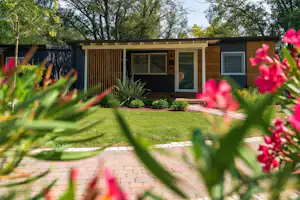 Bungalow mit Terrasse und Sitzbereich, umgeben von Rasen und Büschen, im Hintergrund Bäume und ein Zaun am Campingplatz.