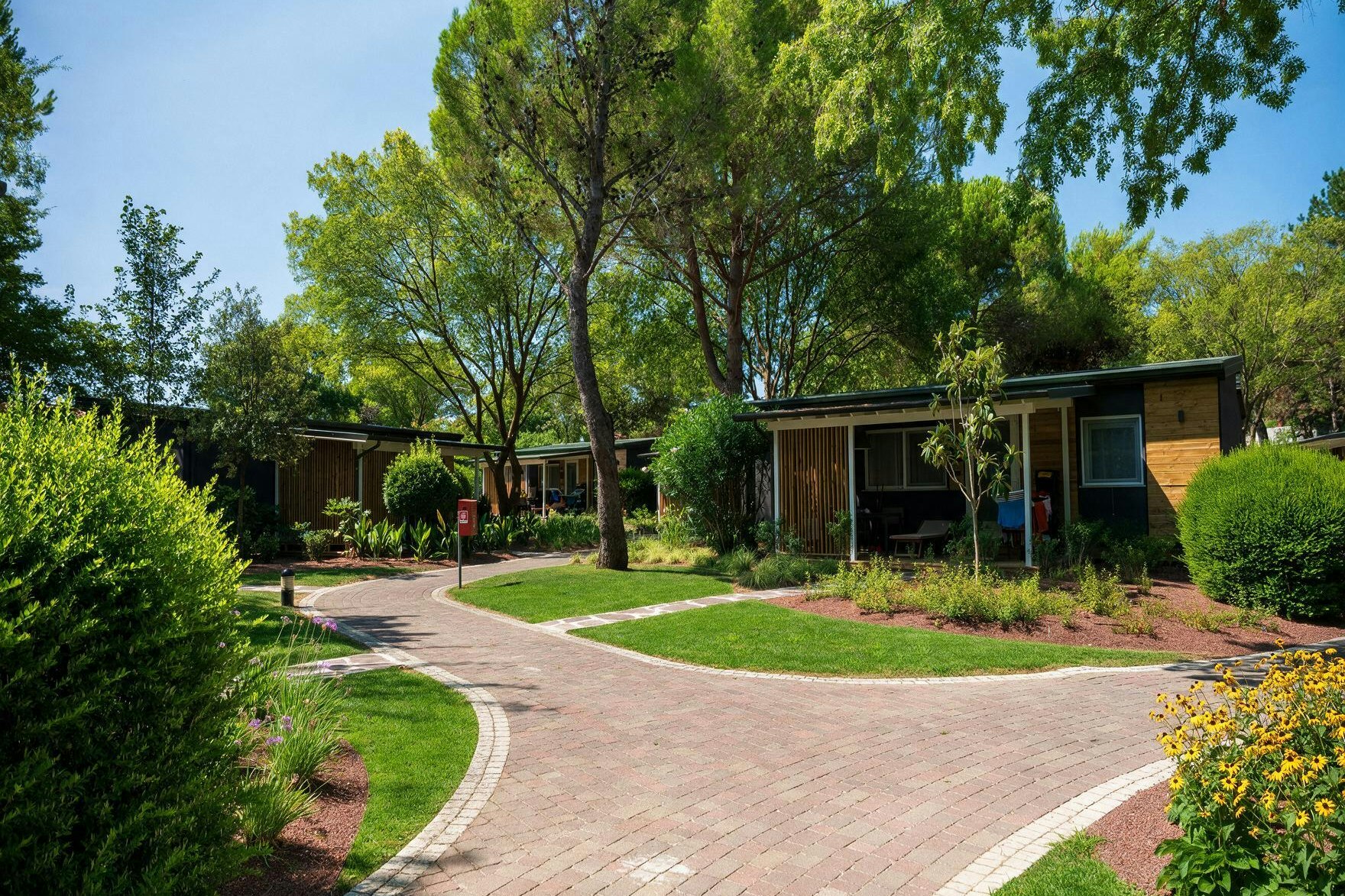 Reihen von Holzbungalows auf einem Campingplatz, mit gepflastertem Weg, Rasenflächen und Bäumen im Hintergrund.