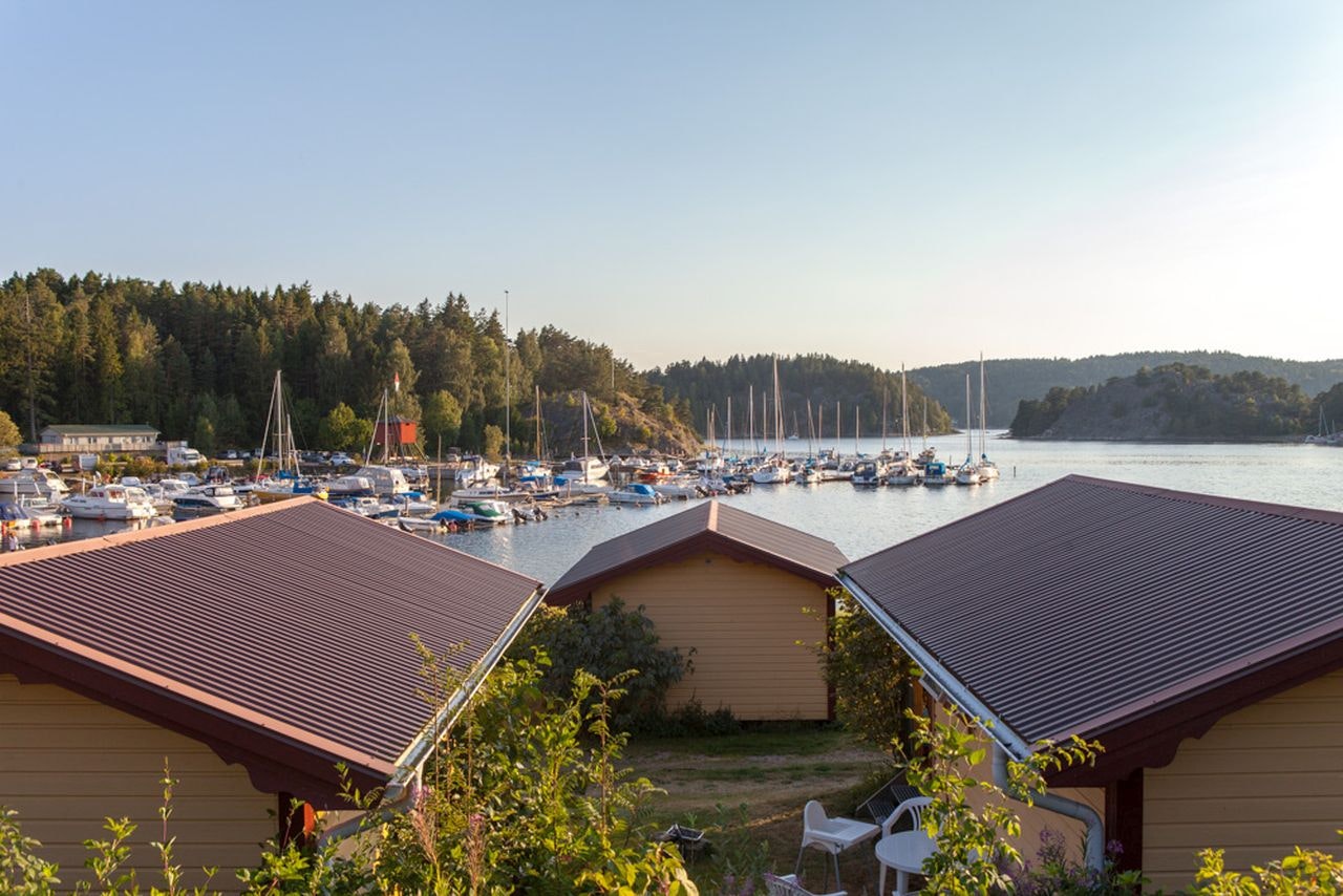 Bungalows mit braunen Dächern am Campingplatz, im Hintergrund viele Boote im See und bewaldete Uferhänge.