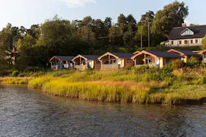 Campingplatz am Fluss mit mehreren Ferienhäusern und kleinen Veranden am Ufer, umgeben von Bäumen und hohem Gras.