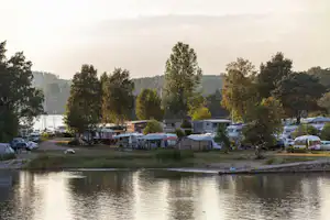 Campingplatz am See mit Wohnwagen und Mobilheimen am Ufer, Bäume und Boote im Hintergrund.