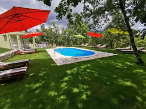 Gepflegter Campinggarten mit blauem Pool, Sonnenliegen und mehreren Sonnenschirmen unter Bäumen.
