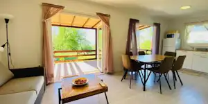 Wohnbereich mit Esstisch und Stühlen, große Fenster mit Balkonzugang, Vorhänge und Küchenzeile in einem Camping-Mobil.