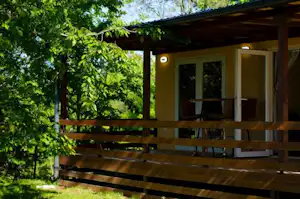 Holzterrasse am Campingunterkunftsgebäude mit Geländer, großen Fenstern und Blick in den grünen Garten mit Bäumen.