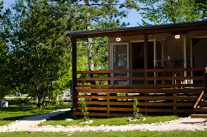 Holzbungalow mit Terrasse und Geländer, Sitzbereich und Weg zum Eingang im Grünen mit Bäumen im Hintergrund.