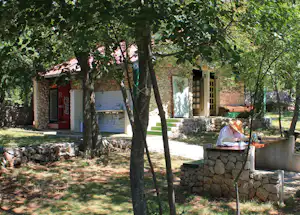 Steinmauer und Terrasse am Campingplatz mit Bäumen im Vordergrund, daneben ein Gebäude mit Eingang und Fenster.