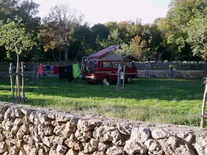 Rotes Wohnmobil auf einem Campingplatz auf einer Wiese, mit Steinmauer, Zaun und Bäumen im Hintergrund.