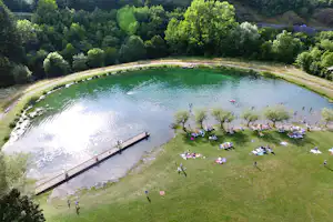 Großer Camping-See mit Schwimmzone, Liegewiese mit Sonnenschirmen und vielen Badegästen, umgeben von Bäumen und Uferwegen.
