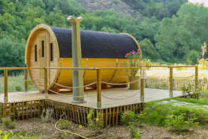Fass-Zelt mit Holzterrasse, Eingangstür und Fenster, davor Hängematte und Blumen im grünen Tal mit Bäumen.