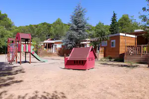Spielplatz mit Rutsche und Klettergerüst auf einem Kiesplatz, im Hintergrund Holzgebäude und Bäume.