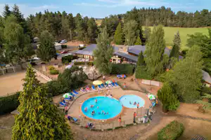 Blick auf Campinganlage mit großem Schwimmbecken, Liegen und Gebäuden, umgeben von Bäumen und Wiesen.