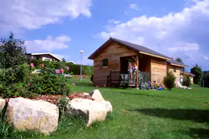 Holzhaus im Campinggarten mit Terrasse, Fahrrädern und gepflegtem Rasen, im Hintergrund weitere Ferienhäuser.