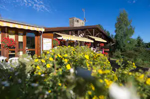 Terrasse eines Camping-Restaurants mit Sonnenschirmen, Holzdach und Blumen im Vordergrund vor einem Gebäude mit Kamin.