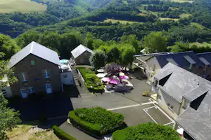 Blick auf einen Campinghof mit Steinhäusern, Terrasse mit Tischen und Sonnenschirmen sowie gepflegten Büschen und weitem Grünland im Tal.