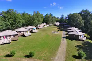 Reihe von Bungalows mit Terrassen und Garten auf einem Campingplatz, dazwischen ein Weg und Tische, umgeben von Bäumen.