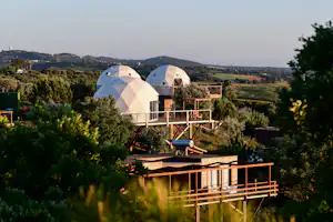 Geodome-Zelte auf einer Holzterrasse, umgeben von Büschen und Feldern, im Hintergrund Hügel und Gebäude.