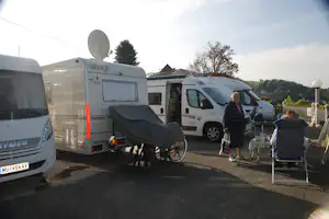 Wohnmobile auf dem Campingstellplatz, mit Markise, Fahrrädern und Stühlen am Weg vor einem Gebäude.