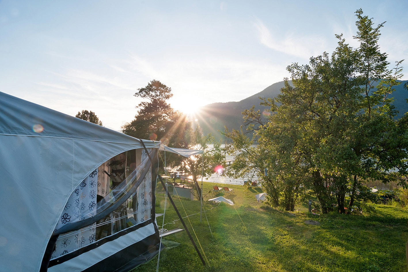 Zelt auf einem Campingplatz am grünen Hang, mit Bäumen, Wiese und Blick auf Berge in der Ferne.