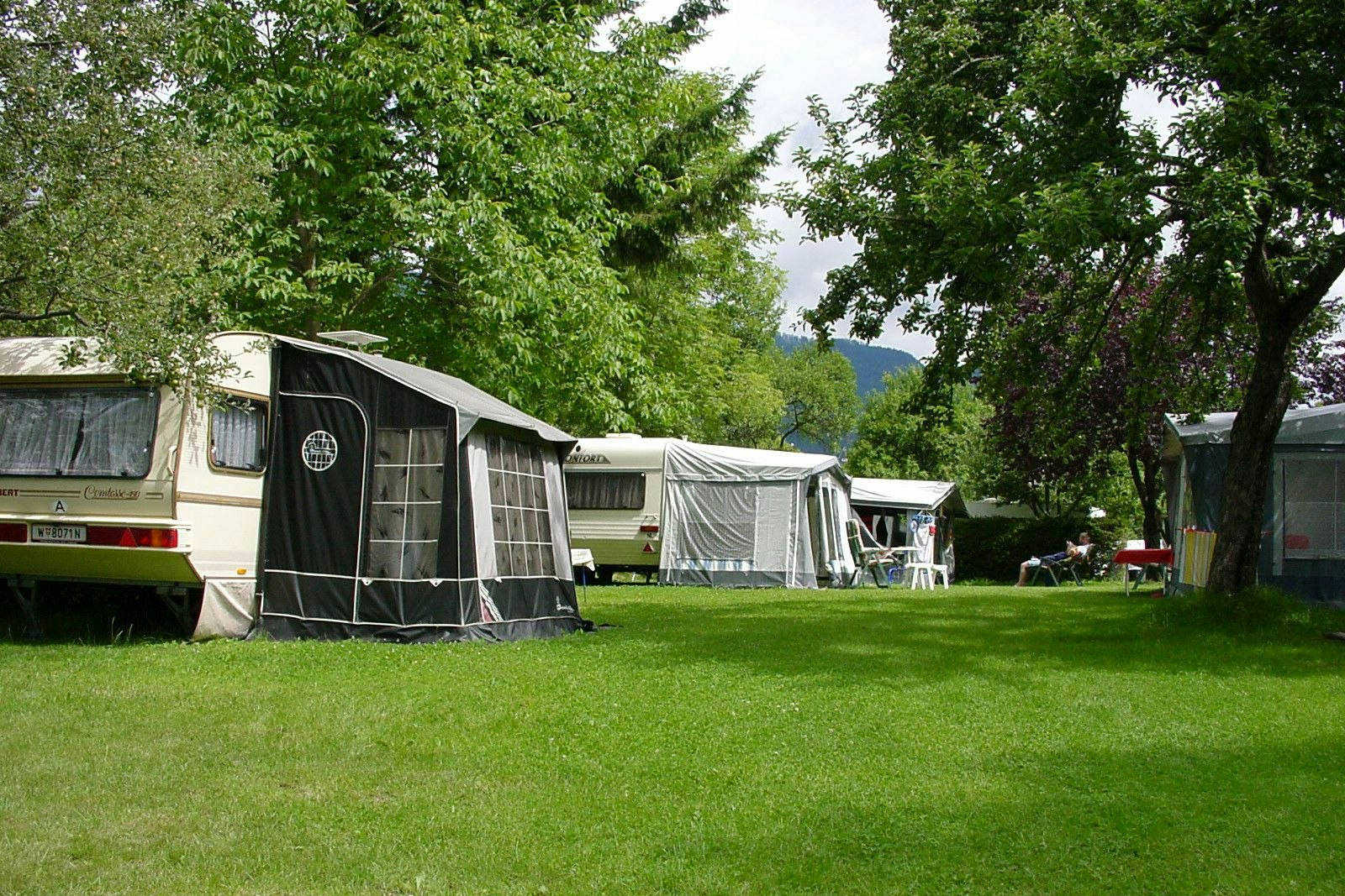 Wohnwagen mit Vorzelt und Markise auf einem grünen Campingplatz, umgeben von Bäumen und weiteren Zelten.