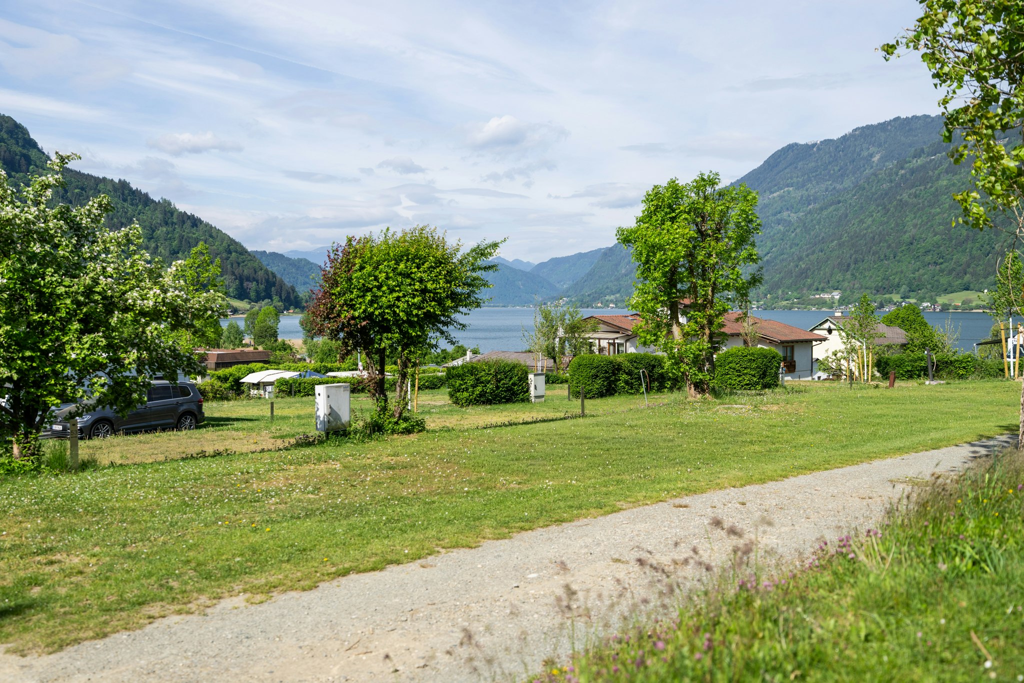 Campingplatz am See mit Rasen, Bäumen und Wohnhäusern, im Hintergrund Berge und ruhiges Wasser.
