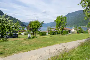 Campingplatz am See mit Rasen, Bäumen und Wohnhäusern, im Hintergrund Berge und ruhiges Wasser.
