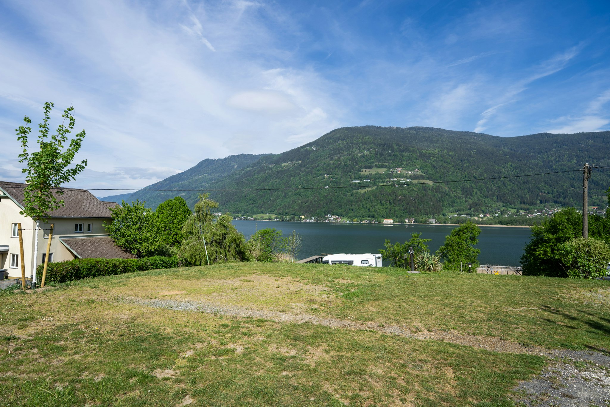 Campingplatz am See mit Blick auf bewaldete Berge, Rasenfläche, Wohnwagen und Gebäude am Ufer.