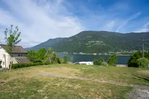 Campingplatz am See mit Blick auf bewaldete Berge, Rasenfläche, Wohnwagen und Gebäude am Ufer.