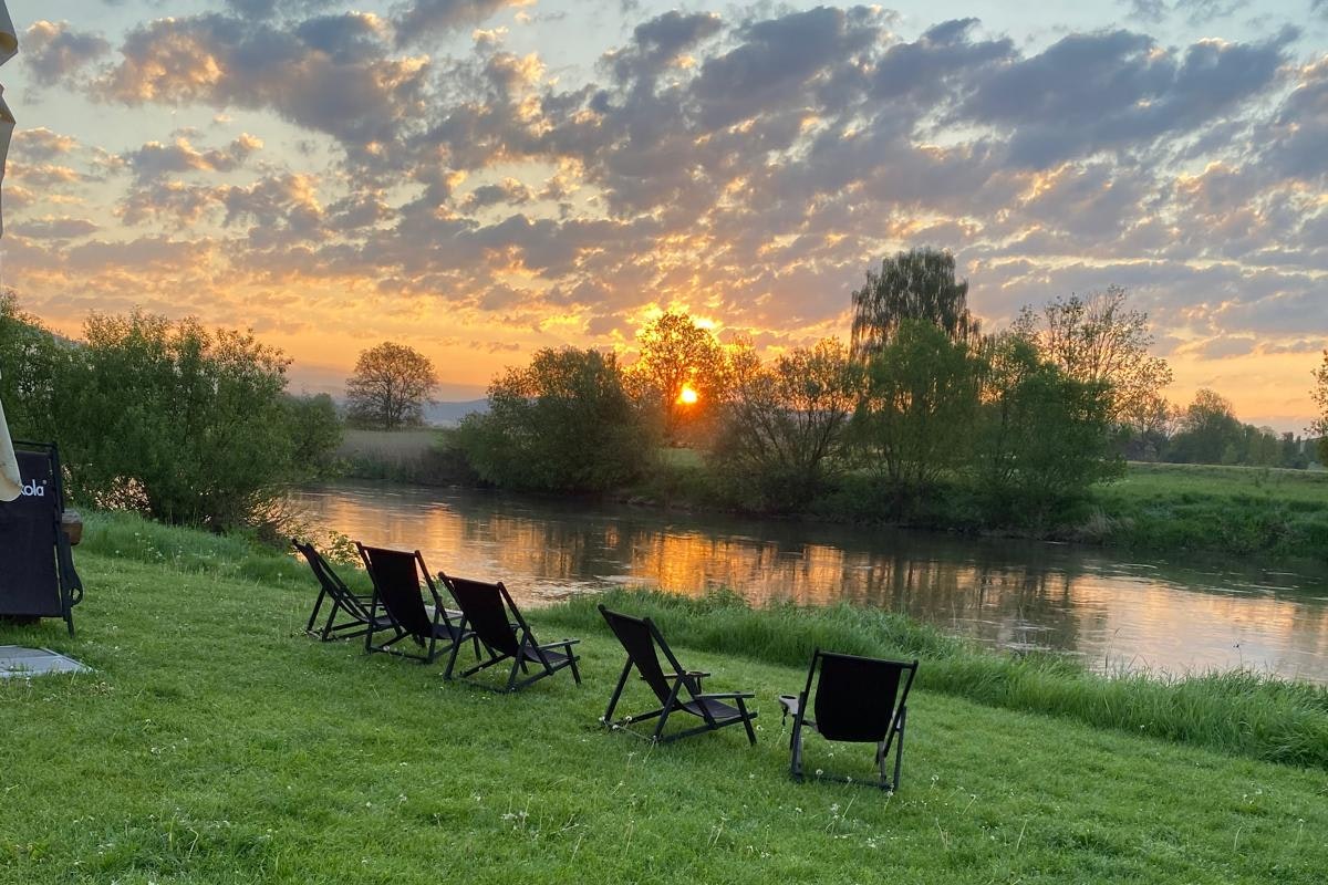 Grüner Campingplatz am Fluss mit Liegestühlen auf dem Rasen, Baumreihe und Sonnenuntergang am Himmel.