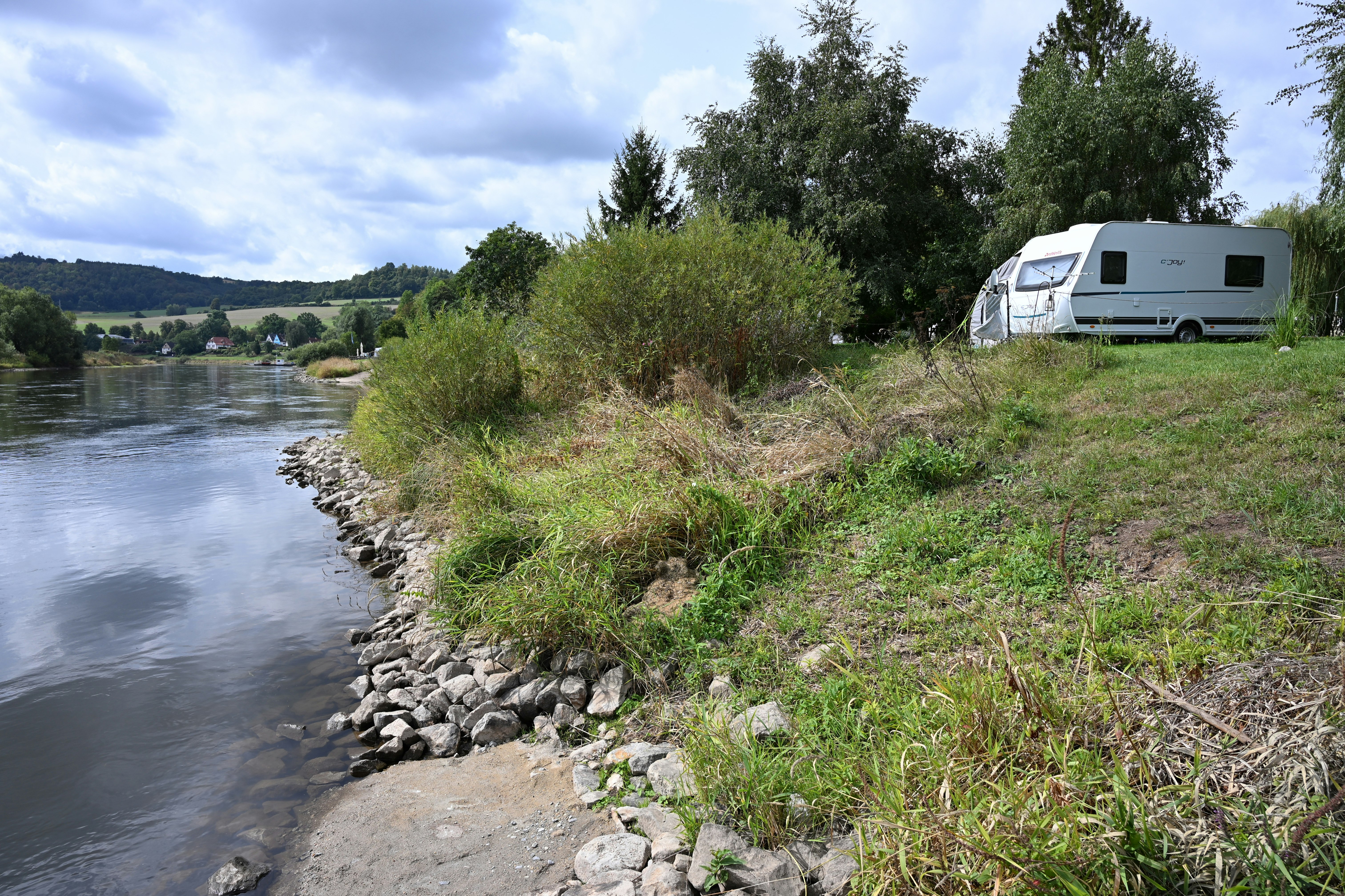 Wohnwagen am Flussufer auf einem Campingplatz, mit Grasfläche, Bäumen und einem befestigten Steinsaum am Wasser.