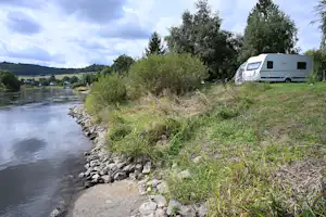 Wohnwagen am Flussufer auf einem Campingplatz, mit Grasfläche, Bäumen und einem befestigten Steinsaum am Wasser.
