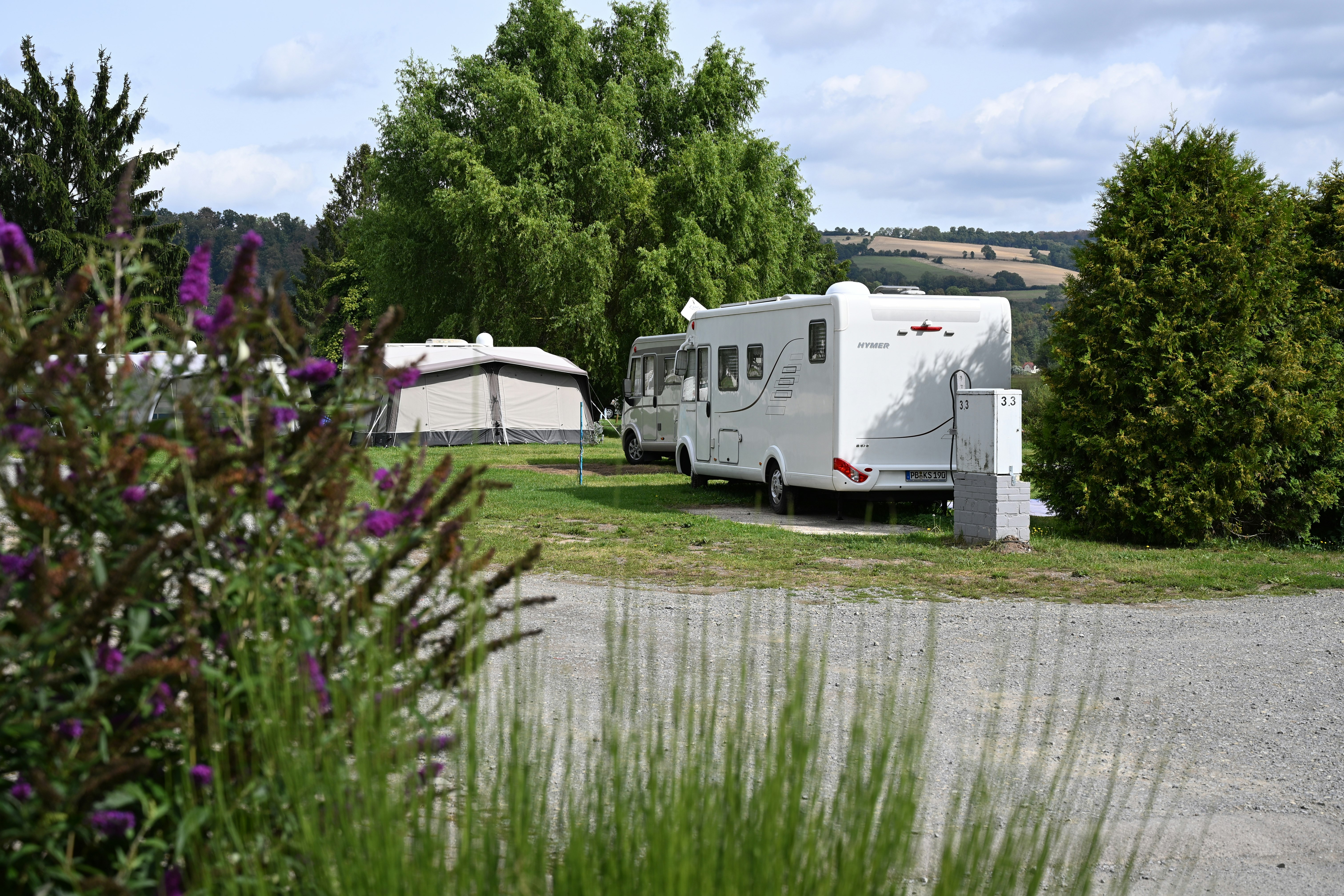 Wohnmobil auf einem Campingplatz neben einem kleinen Zelt, umgeben von Gras, Büschen und Bäumen am Weg.