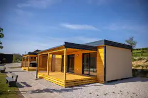 Modernes Holz- und Wohnmobilhaus mit großer Holzterrasse, Glasfront und Schiebetüren auf Kiesstellplatz, daneben Sitzbank.