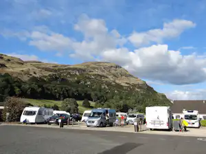Wohnmobile und Campingfahrzeuge auf einem Stellplatz, im Hintergrund bewaldete Berge und ein Parkplatz mit Schranken.