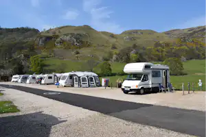Wohnmobile auf einem Campingplatz am Straßenrand, mit Stellplätzen, Absperrpfosten und Blick auf grüne Hügel im Tal.