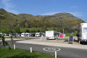 Wohnmobile auf einem Stellplatz am Campingplatz, mit Schildern, Absperrpfosten und Blick auf grüne Hügel und Berge.