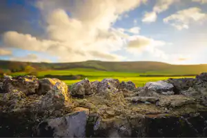 Steinmauer mit Felsbrocken im Vordergrund, dahinter grüne Hügel und ein Himmel mit Wolken über einer Campinglandschaft.