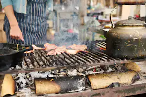 Grill mit Fleisch auf dem Rost, Funken und Rauch beim Kochen am Campingplatz, daneben Pfanne und Holzkohle.