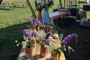 Blumenstrauß auf einem Holztisch unter einem Zeltvordach, im Hintergrund weitere Zelte und eine grüne Wiese.