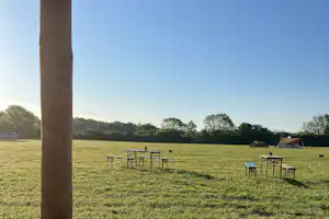 Campingwiese mit Holztisch und Stühlen, im Hintergrund Zelte und eine weite Landschaft unter klarem Himmel.
