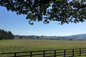 Weite Wiese mit Zaun im Vordergrund, dahinter Felder und Hügel unter klarem Himmel, umgeben von Bäumen.