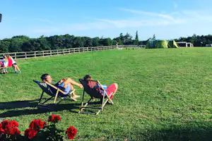 Zwei Camping-Liegen auf einer grünen Wiese, im Hintergrund ein Zelt und ein weißer Zaun.