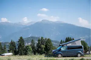 Camping-Van auf einer Wiese mit geöffnetem Dach, im Hintergrund Tannen und ein weites Bergpanorama.
