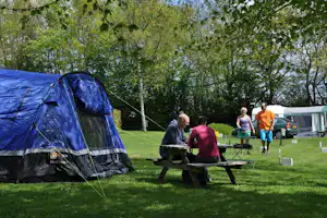 Blaues Campingzelt neben einem Holztisch mit Grill auf dem Rasen, im Hintergrund weitere Zelte und Wohnwagen.